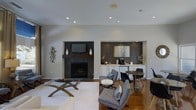 This single-floor space on Lake Lynn Drive in Raleigh, North Carolina, showcases a contemporary interior design with a cohesive, open layout. The main living area centers around a striking fireplace framed by dark tile surround and a rich wood mantel, paired with a recessed media niche that creates a polished, built-in aesthetic. Recessed lighting throughout the smooth white ceiling enhances the bright, airy atmosphere of the room.

The living area features warm hardwood flooring contrasted by a large white area rug, anchoring a seating arrangement of modern upholstered chairs in soft gray tones. A glass-top coffee table with a sculptural wood base adds an artistic touch to the space.

Adjacent to the living area, a dedicated wet bar niche provides functional convenience, complete with white cabinetry, a light countertop, and a dark textured accent wall backdrop. A small bistro-style table and chairs in this zone create an intimate setting for casual use. Decorative wall art and sculptural accents throughout the space contribute to its refined, contemporary character.