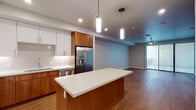 This single-floor residence showcases a contemporary design characterized by clean lines, warm wood-toned finishes, and an open, connected layout. The kitchen serves as the heart of the space, featuring a central island with a white countertop that provides both workspace and casual seating. Cabinetry combines rich walnut-toned lower cabinets with crisp white upper cabinets, complemented by a white tile backsplash and stainless steel appliances, including a French door refrigerator with an ice and water dispenser, a dishwasher, and a range. Pendant lighting above the island adds a modern accent. The kitchen flows seamlessly into a generously sized living area finished with hardwood-style flooring and a ceiling fan, with sliding glass doors opening to an outdoor balcony. Bedrooms are well-proportioned with neutral walls, wood-style flooring, and ceiling-mounted lighting. The bathrooms continue the warm aesthetic, with the primary bath offering a dual-sink vanity topped with white countertops, warm wood cabinetry, and a large wall-spanning mirror. A secondary bath mirrors the clean, minimal style. Throughout, recessed lighting, neutral gray walls, and consistent flooring create a cohesive, modern ambiance.