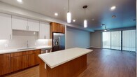 This single-floor residence showcases a contemporary design characterized by clean lines, warm wood-toned finishes, and an open, connected layout. The kitchen serves as the heart of the space, featuring a central island with a white countertop that provides both workspace and casual seating. Cabinetry combines rich walnut-toned lower cabinets with crisp white upper cabinets, complemented by a white tile backsplash and stainless steel appliances, including a French door refrigerator with an ice and water dispenser, a dishwasher, and a range. Pendant lighting above the island adds a modern accent. The kitchen flows seamlessly into a generously sized living area finished with hardwood-style flooring and a ceiling fan, with sliding glass doors opening to an outdoor balcony. Bedrooms are well-proportioned with neutral walls, wood-style flooring, and ceiling-mounted lighting. The bathrooms continue the warm aesthetic, with the primary bath offering a dual-sink vanity topped with white countertops, warm wood cabinetry, and a large wall-spanning mirror. A secondary bath mirrors the clean, minimal style. Throughout, recessed lighting, neutral gray walls, and consistent flooring create a cohesive, modern ambiance.