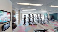 This single-floor fitness center offers a well-appointed exercise environment designed to support a variety of workout routines. The space is outfitted with a row of cardio machines, including ellipticals and step trainers, each equipped with individual monitors for entertainment or performance tracking. A large wall-to-wall mirror runs along one side of the room, effectively doubling the sense of space and allowing users to monitor their form during exercise. Natural light filters in through windows at the far end, contributing to an open and energizing atmosphere. Recessed panel lighting in the textured ceiling provides even illumination throughout. The flooring features a bold, multi-colored patchwork carpet in shades of pink, blue, and tan, adding a vibrant and contemporary design element to the space. Stability and medicine balls are neatly stored near a support column, indicating a dedicated area for functional and core training. A teal accent wall is partially visible, adding a pop of color and modern flair. Overall, the fitness center presents a clean, functional, and visually dynamic environment suited for both cardio and strength-focused workouts.