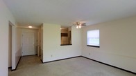 This single-floor space features an open living area finished with wall-to-wall carpet and neutral cream-colored walls, creating a clean, understated atmosphere. A ceiling fan with integrated lighting is centrally positioned overhead, providing both airflow and ambient illumination. A window along one wall admits natural light, brightening the space throughout the day.

A partial dividing wall separates the living area from the kitchen, which is accessible via a pass-through opening—a practical architectural detail that maintains visual connectivity between the two spaces while defining separate functional zones. The kitchen area, visible through the pass-through, features upper cabinetry and appears to include a refrigerator.

Toward the entry, the flooring transitions from carpet to tile, demarcating the entryway from the main living space. A door at the far end leads to additional areas of the unit, including a hallway that connects to further rooms. Dark baseboards provide a subtle contrast against the light walls throughout.

The textured ceiling adds a traditional finish to the overall interior. The layout is practical and functional, with clearly defined zones for living, dining, and kitchen activity within a compact, single-level footprint.