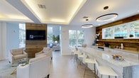 This single-floor clubhouse in Diamond offers a beautifully curated space that seamlessly blends contemporary design with warm, rustic elements. The open-concept layout is anchored by striking reclaimed wood columns and exposed ceiling beams that contrast handsomely against crisp white walls and ceilings, creating a welcoming yet sophisticated atmosphere throughout.

The main gathering area features a generous bar counter finished in luxurious marble-look stone, lined with sleek white bar stools and framed by a reclaimed wood accent wall fitted with display shelving. Overhead, modern circular pendant lights provide a refined focal point, while a tray ceiling with integrated ambient lighting adds depth and warmth to the space.

A comfortable lounge area outfitted with plush neutral-toned armchairs provides an inviting setting for relaxation or socializing, complemented by a large flat-screen television mounted on a matching wood feature wall. Light-toned tile flooring flows continuously throughout, enhancing the sense of openness and cohesion.

Large windows on multiple walls flood the interior with natural light and offer views of the lush outdoor surroundings and pool area beyond the glass entry doors. The overall ambiance is airy, polished, and thoughtfully designed to encourage community and leisure.