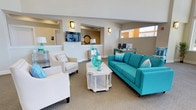 Located at 100 Isabelle Isle in Dover, Delaware, this single-floor community space offers a thoughtfully designed environment that blends comfort and functionality. The lounge area serves as the heart of the space, featuring a vibrant teal sofa and coordinating accent chairs arranged around an ornate white coffee table, creating an inviting gathering spot. The coastal-inspired color palette — combining aqua tones with neutral beige walls and carpeting — lends the interior a fresh, relaxed ambiance. Wall-mounted sconces provide warm accent lighting, complementing the natural light that filters in through clerestory windows positioned along the upper walls. Just beyond the lounge, an open passageway leads to a reception or greeting area, complete with a wall-mounted television and a round decorative mirror, adding a refined touch to the transition space. To the right of the lounge, a dedicated business center is equipped with desktop computers and standing workstations, offering residents a practical area for everyday tasks. The open-concept layout promotes easy flow between zones while maintaining a sense of purpose in each area. Overall, the space strikes a balance between a welcoming social atmosphere and practical amenities, making it a versatile and well-appointed community hub.