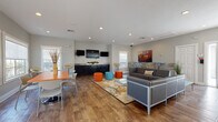 Located at 1700 Woodbury Rd in Orlando, Florida, this single-story community clubhouse offers a bright, open-plan gathering space designed for socializing and relaxation. Rich hardwood-style flooring flows throughout, complementing the neutral gray walls and recessed lighting that create a clean, contemporary atmosphere.

The main lounge area features a sleek gray sectional sofa arrangement centered on a patterned area rug, accented by vibrant orange and teal cylindrical ottomans that add playful pops of color. A media wall with multiple wall-mounted flat-screen televisions sits above dark cabinetry with granite countertops, making it ideal for group viewing. Colorful artwork, including a bold cityscape canvas, adds visual interest to the walls.

A dedicated dining zone is furnished with a bold orange communal table and modern chairs, bathed in natural light from large windows with views of the surrounding landscaping. Bistro-height bar tables and swivel stools provide additional casual seating throughout.

Glass entry doors open to an outdoor area, enhancing the flow between indoor and outdoor spaces. The room also includes a foosball table for recreation. Ample window placement ensures the entire space is filled with Florida sunshine, fostering a welcoming and energetic environment suitable for community events and everyday gatherings.