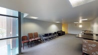 The 2nd Floor Lounge at 209 is a versatile, open-plan space designed to accommodate both collaborative work and casual gathering. The room features a sloped ceiling with recessed skylights that introduce natural light throughout the day, complementing the flush-mounted ceiling fixtures that ensure consistent illumination across the space.

The flooring throughout is a neutral grey carpet, lending a subdued, functional aesthetic to the room. Along one wall, a mix of seating options is arranged, including upholstered chairs of varying styles and a long, foldable work table flanked by rolling task chairs, offering a practical setup for meetings or group work sessions. Toward the far end of the room, a dark upholstered sofa provides a more relaxed seating area.

The walls are painted in soft, muted tones — primarily light grey with a warm yellow accent visible in the adjoining area — contributing to an understated, neutral ambiance. The open layout and generous floor area allow for flexible furniture arrangement to suit a variety of uses. Overall, the lounge presents a functional and adaptable environment suited for both focused work and informal social interaction.