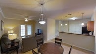 This single-floor residence features an open-concept layout that seamlessly connects the dining area, living room, and kitchen into one cohesive living space. The dining area is anchored by a round dark-wood table with matching chairs, positioned beneath a contemporary pendant light fixture that draws the eye upward. Crown molding lines the ceiling, adding a refined architectural detail to the space.

The living room area is carpeted and receives natural light through a set of French doors, which also provide an entry point and enhance the room's airy feel. A ceiling fan is mounted overhead, contributing both to comfort and casual style. The space is furnished with a neutral-toned sofa and includes a media console with a flat-screen television.

Transitioning toward the back of the space, a raised breakfast bar counter separates the kitchen from the open living area. Two additional pendant lights hang above the bar, providing focused task lighting. The kitchen features white cabinetry and light-colored walls, maintaining the home's consistent neutral palette. The overall ambiance is bright, functional, and well-suited for everyday living and entertaining.