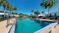 Located at 303 Sutton Cir in Daytona Beach, Florida, this outdoor community pool area offers a resort-style recreational experience in a sun-drenched Florida setting. The pool itself is generously sized, featuring a deep end that reaches 8 feet 6 inches, making it suitable for both swimmers and those seeking a refreshing dip. The water is crystal clear with a striking turquoise hue, framed by classic blue tile accents along the pool's edge that add a polished, decorative touch.

Surrounding the pool is a wide, open concrete sun deck that provides ample space for relaxation and outdoor gatherings. The deck is well-furnished with rows of lounge chairs and seating arrangements, ideal for sunbathing or socializing in the warm Florida climate. A secondary, shallower pool or spa area is also visible, connected to the main pool, offering additional options for leisure.

Tall, mature palm trees line the perimeter of the pool area, creating a tropical ambiance and providing natural shade throughout the day. The space is enclosed by a black iron security fence, ensuring both safety and privacy. The surrounding residential buildings, finished in white and light gray exteriors, are visible in the background, complementing the clean, bright aesthetic of the overall environment. Under the consistently clear blue Florida skies, this pool area serves as a standout communal amenity.