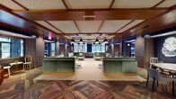 This thoughtfully designed single-floor amenity space blends industrial character with refined comfort across several distinct zones. The centerpiece is a spacious lounge area defined by exposed brick columns, coffered wood-beamed ceilings, and striking herringbone-patterned hardwood floors. Plush green velvet sectional sofas are arranged symmetrically throughout, paired with marble-top coffee tables and anchored by bold pendant lighting suspended from the ceiling. Large windows along the perimeter flood the space with natural light and offer views of an adjacent outdoor courtyard, which is accessible through glass double doors framed by decorative terracotta-toned patterned tile flooring. A gallery wall with curated artwork and a built-in banquette seating area add warmth and personality to one corridor. The space also includes a dining and kitchenette nook featuring dark walls, patterned cement tile floors, and a light wood countertop with under-counter appliances — ideal for casual gatherings. A service area showcases a bold green-and-white scallop tile accent wall alongside intricate mosaic floor tiles, adding a layer of artistic detail. Throughout, the design balances eclectic tile work, natural materials, and curated furnishings to create an inviting, multi-functional communal environment.