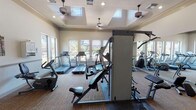 This fitness center offers a comprehensive and well-organized workout environment designed to support a wide range of exercise routines. The space is flooded with natural light through large multi-pane windows that line the walls, offering pleasant views of the surrounding outdoor landscaping and community. Tray ceilings with skylights in the main workout area further enhance the bright, open atmosphere, while ceiling fans provide comfortable air circulation throughout.

The cardio section is well-appointed, featuring multiple Cybex treadmills, elliptical machines, and stationary and recumbent bikes, all positioned to take advantage of the outdoor views. Strength training is supported by a Precor multi-station cable machine along with additional resistance equipment, providing options for a full-body workout. Full-wall mirrors on one side of the room expand the sense of space and allow users to monitor their form during exercise.

The flooring throughout is a durable, textured carpet with protective rubber mats beneath each piece of equipment. Wall-mounted televisions add an entertainment option for cardio sessions. Informational bulletin boards and posted notices are visible near the entry area. The fitness center is accessible via a glass-panel door with an adjacent viewing window, and the overall design maintains a clean, neutral aesthetic with warm beige tones that create an inviting and motivating workout environment.