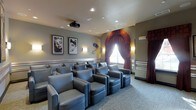 This private screening room is thoughtfully designed to deliver an authentic cinematic experience. The space is arranged with rows of plush, blue-gray leather lounge chairs positioned in a tiered layout, ensuring clear sightlines to the screen from every seat. A ceiling-mounted projector anchors the room's purpose, while recessed lighting and wall-mounted sconces provide warm, ambient illumination that complements the theater atmosphere.

The décor takes clear inspiration from the golden age of Hollywood. Framed black-and-white portraits of iconic film legends adorn the walls, adding a classic, glamorous character to the room. A decorative arched alcove dressed with rich burgundy drapes serves as a striking focal point, evoking the grandeur of a traditional cinema entrance. Coordinating curtains frame a window, reinforcing the cohesive, vintage aesthetic throughout.

The walls are finished in a soft, neutral gray tone accented with a decorative chair rail, while patterned carpeting in muted tones adds texture underfoot. Built-in side tables and armrests enhance guest comfort. The overall ambiance is intimate yet polished, making this screening room an ideal venue for private movie viewings or entertainment gatherings.