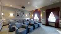 This private screening room is thoughtfully designed to deliver an authentic cinematic experience. The space is arranged with rows of plush, blue-gray leather lounge chairs positioned in a tiered layout, ensuring clear sightlines to the screen from every seat. A ceiling-mounted projector anchors the room's purpose, while recessed lighting and wall-mounted sconces provide warm, ambient illumination that complements the theater atmosphere.

The décor takes clear inspiration from the golden age of Hollywood. Framed black-and-white portraits of iconic film legends adorn the walls, adding a classic, glamorous character to the room. A decorative arched alcove dressed with rich burgundy drapes serves as a striking focal point, evoking the grandeur of a traditional cinema entrance. Coordinating curtains frame a window, reinforcing the cohesive, vintage aesthetic throughout.

The walls are finished in a soft, neutral gray tone accented with a decorative chair rail, while patterned carpeting in muted tones adds texture underfoot. Built-in side tables and armrests enhance guest comfort. The overall ambiance is intimate yet polished, making this screening room an ideal venue for private movie viewings or entertainment gatherings.