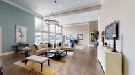 Located at 2402 N Ben Wilson St in Victoria, Texas, this single-floor community clubhouse delivers an impressive blend of refined design and functional amenities. The centerpiece of the space is a grand open lounge area distinguished by soaring ceilings, floor-to-ceiling windows, and an elegant wrought-iron chandelier that anchors the room. Rich herringbone-patterned hardwood flooring runs throughout, complementing a neutral color palette accented by soft teal walls and crisp white trim. Oversized windows flood the interiors with abundant natural light and offer views of the surrounding landscaped outdoor areas, including an accessible patio and pool deck. The lounge is thoughtfully furnished with plush neutral sofas, accent pillows, and a colorful area rug, creating an inviting gathering space. A dedicated game room features a full-size shuffleboard table, wall-mounted televisions, and seating for leisure and entertainment, with direct access to the outdoor terrace. The kitchen is equipped with white shaker cabinetry, dark countertops, a marble-veined island, mosaic tile backsplash, and modern stainless steel appliances — ideal for hosting events. Additional areas include a hallway corridor with display storage and signage directing to a 24-hour fitness center, completing this well-rounded community facility.