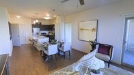 This single-floor residence showcases an open-concept layout that seamlessly connects the living, dining, and kitchen areas into one cohesive, functional space. The hardwood-style flooring runs throughout, providing a warm and consistent foundation that ties each zone together visually.

The kitchen is appointed with dark cabinetry that creates a striking contrast against lighter countertops and stainless steel appliances. A central island with bar seating offers additional prep space and casual dining options, while pendant lighting above adds both task illumination and stylish detail.

Adjacent to the kitchen, the dining area accommodates a glass-top table surrounded by upholstered chairs, making it well-suited for both everyday meals and entertaining. A large-scale abstract artwork in warm golden tones anchors the nearby wall, adding a refined artistic touch.

The living area flows naturally from the dining space, featuring a ceiling fan overhead and room for comfortable seating arrangements. Neutral wall tones throughout keep the space feeling bright and versatile, allowing the furnishings and décor to take center stage. Overall, the layout emphasizes practicality and modern design in equal measure.