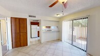 This single-floor unit offers a functional and efficient layout with a cohesive design throughout. The main living and dining area is bright and open, featuring large sliding glass doors fitted with vertical blinds that invite natural light and provide access to an outdoor space with views of surrounding greenery. A ceiling fan with a light fixture keeps the space comfortable, while light large-format tile flooring extends across the common areas, giving the space a clean, airy feel.

The kitchen is accessible directly from the living area and is also connected via a convenient pass-through counter, adding an open, connected quality to the layout. It is equipped with warm wood-toned cabinetry, a white electric range with an overhead ventilation hood, a full-size refrigerator, and ample counter space for meal preparation. A window with vertical blinds provides natural light and outdoor views.

A bedroom features soft carpet flooring and a generously sized walk-in closet with built-in shelving and tile flooring — offering practical storage solutions. The bathroom is finished in clean white tones and includes a full-size soaking tub with a shower combination, a toilet, and towel bars. Textured ceilings are present throughout, and the overall palette of neutral whites and warm wood accents creates a simple, functional living environment.