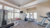 This fitness center is designed to provide a comprehensive workout environment within a bright, open single-floor layout. The space is characterized by warm beige walls, tray ceilings with crown molding details, and durable patterned carpet flooring suited for high-traffic exercise use. Large, multi-pane windows line the walls throughout, flooding the interior with natural light and offering views of the surrounding greenery, creating an inviting and energizing atmosphere.

The facility is thoughtfully divided into distinct workout zones. The cardio area accommodates multiple treadmills and elliptical machines, positioned to take full advantage of the window-facing views. A dedicated strength training section houses a multi-station cable machine, weight benches, and additional resistance equipment, providing options for a full-body workout. Stability balls add a functional fitness element to the space.

Wall-mounted flat-screen televisions are positioned throughout for entertainment during workouts. Full-length mirrors along one wall enhance the sense of space while allowing users to monitor their form. The fitness center also features a mirrored glass door entry, recessed lighting, and proper ventilation, ensuring a comfortable and well-maintained exercise environment. Overall, the space strikes a balance between functionality and a welcoming aesthetic.