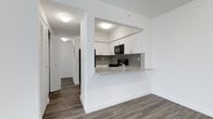 This single-floor unit offers a clean, modern aesthetic defined by a cohesive design language carried throughout the space. The open-concept kitchen is a central highlight, featuring crisp white cabinetry that extends to the ceiling, granite-look countertops, and a tasteful mosaic tile backsplash that adds visual texture. Black appliances, including a range and over-the-range microwave, provide a striking contrast against the bright white finishes. A peninsula counter with an overhang serves as a practical breakfast bar, seamlessly connecting the kitchen to the main living area and enhancing the sense of open flow. The flooring throughout is a warm-toned, wood-style laminate that adds durability and a welcoming character to every room. The hallway, visible from the main living area, leads to additional rooms and features a louvered closet door, suggesting generous storage options. Flush-mount ceiling fixtures provide consistent, even lighting across the space. The walls are finished in a clean, neutral white, amplifying the brightness and creating a versatile backdrop. Overall, the unit presents a well-maintained, move-in-ready interior with a contemporary feel.