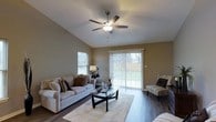 This single-floor residence offers a welcoming and well-appointed living space designed for both comfort and functionality. The living room serves as the heart of the home, characterized by soaring vaulted ceilings that lend an airy, open feel to the space. A ceiling fan with an integrated light fixture is centrally mounted, providing both circulation and illumination. Rich, dark hardwood flooring runs throughout, adding warmth and a polished aesthetic to the interior. Natural light flows generously into the room through a large sliding glass door fitted with vertical blinds, offering a direct view of the outdoor yard area, as well as an additional side window. The neutral wall tones complement the light-colored furnishings, creating a calm and cohesive atmosphere. The living area comfortably accommodates a full seating arrangement, with ample space for relaxed gatherings. The overall ambiance is bright, clean, and inviting, with thoughtful architectural details — particularly the vaulted ceiling line — giving the space a distinctive character. The seamless connection between the interior and the backyard through the sliding door enhances the sense of openness and provides convenient outdoor access.