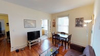 This single-floor residence combines its living and dining areas into one cohesive, open space anchored by warm hardwood floors that extend throughout. The living area accommodates a flat-screen television on a light wood stand alongside a glass-top coffee table, while a dark wood dining set with four chairs sits beneath a bright window fitted with white blinds, allowing ample natural light to fill the room. The walls are painted in a soft, neutral tone, contributing to a clean and airy atmosphere. A doorway visible from the main living space leads into what appears to be a kitchen, featuring light wood cabinetry and additional window light, suggesting a functional and well-lit cooking area. Baseboard heating units line the walls, indicating a practical heating setup. The space is decorated simply, with a wall map and framed artwork adding character without overwhelming the room. Overall, the unit presents a straightforward, livable layout that balances the living and dining functions efficiently within a compact but comfortable footprint.