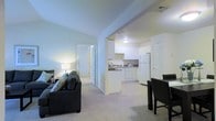 This single-floor residence showcases an open-concept design that seamlessly connects the living, dining, and kitchen areas into one cohesive space. The living area is carpeted and comfortably accommodates a dark upholstered sofa accented with decorative pillows, complemented by a softly lit floor lamp that adds warmth to the room. A hallway visible from the main living space leads to additional rooms, including what appears to be a bedroom and a full bathroom, providing a clear and practical flow throughout the unit.

The kitchen is positioned in a partially open alcove, distinguished by its white cabinetry, white appliances including a full-size refrigerator, and recessed countertop workspace. A ceiling-mounted light fixture illuminates the kitchen area effectively. Adjacent to the kitchen, a dining area features dark seating, creating a natural transition between cooking and dining spaces. The walls throughout are painted in neutral tones, and the consistent light carpeting across the main living areas ties the space together visually. The layout is efficient, making excellent use of the available floor space while maintaining an open and airy atmosphere.
