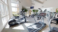 This fitness center is designed to offer a bright, energizing environment for a full range of workouts. The space is defined by soaring vaulted ceilings that create an open, airy atmosphere, complemented by large floor-to-ceiling windows that flood the room with abundant natural light and provide pleasant outdoor views. Multiple ceiling fans are positioned throughout to ensure comfortable airflow during exercise sessions.

The cardio area is well-appointed with a selection of treadmills, elliptical trainers, and other aerobic machines, including Star Trac branded equipment. A wall-mounted flat-screen television is positioned at an optimal viewing angle for those using the cardio equipment. The strength training zone offers a variety of resistance and weight machines, catering to users of all fitness levels.

Lush indoor plants are thoughtfully placed throughout the space, adding a natural, refreshing element to the workout environment. Large mirrors line one wall, enhancing the sense of openness and allowing users to monitor their form. The neutral-toned flooring ties the entire space together, creating a clean and professional aesthetic. Overall, this fitness center provides a welcoming and fully equipped setting for an effective workout experience.