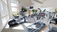 This fitness center is designed to offer a bright, energizing environment for a full range of workouts. The space is defined by soaring vaulted ceilings that create an open, airy atmosphere, complemented by large floor-to-ceiling windows that flood the room with abundant natural light and provide pleasant outdoor views. Multiple ceiling fans are positioned throughout to ensure comfortable airflow during exercise sessions.

The cardio area is well-appointed with a selection of treadmills, elliptical trainers, and other aerobic machines, including Star Trac branded equipment. A wall-mounted flat-screen television is positioned at an optimal viewing angle for those using the cardio equipment. The strength training zone offers a variety of resistance and weight machines, catering to users of all fitness levels.

Lush indoor plants are thoughtfully placed throughout the space, adding a natural, refreshing element to the workout environment. Large mirrors line one wall, enhancing the sense of openness and allowing users to monitor their form. The neutral-toned flooring ties the entire space together, creating a clean and professional aesthetic. Overall, this fitness center provides a welcoming and fully equipped setting for an effective workout experience.