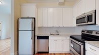 This single-floor space is thoughtfully designed with a clean, modern aesthetic. The kitchen serves as a standout feature, showcasing crisp white shaker-style cabinetry that extends to the ceiling, maximizing storage capacity. Granite countertops with subtle veining provide a durable and elegant work surface, complemented by a stainless steel sink with a pull-out faucet. The kitchen is fully equipped with stainless steel appliances, including a full-size refrigerator, dishwasher, microwave, and range, offering a cohesive and contemporary look. Crown molding along the upper perimeter of the kitchen adds a refined architectural touch, elevating the overall design. Warm hardwood flooring runs underfoot, providing a natural contrast to the bright white cabinetry and light-toned walls. The layout is efficient and functional, making excellent use of the available space. Natural light enhances the airy, open feel of the kitchen, highlighting the clean lines and neutral color palette throughout. Overall, the space presents a polished, move-in-ready environment with quality finishes and practical design elements.