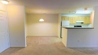 This single-floor space presents an open-concept layout that seamlessly connects the living, dining, and kitchen areas into one cohesive environment. The living and dining area is finished with neutral-toned wall-to-wall carpeting, lending a soft, comfortable feel underfoot. A single dome pendant light hangs centrally, providing a defined focal point for the dining area while illuminating the space effectively. The walls are finished in a clean, light neutral tone that enhances the brightness of the room.

Adjacent to the living area, the kitchen is separated by a raised breakfast bar counter, creating a natural boundary while maintaining visual connectivity between spaces. The kitchen features light wood cabinetry, a white refrigerator, and dark countertops, offering a functional layout. Overhead lighting in the kitchen ensures the workspace remains well-lit. The raised counter provides additional seating or prep space and adds a practical element to the overall design.

The open flow between rooms contributes to an airy, uncluttered ambiance, making the space feel welcoming and versatile. The neutral palette throughout allows for flexible interior styling.
