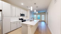 Located at 1350 W Taylor St in Sherman, TX, this thoughtfully designed one-bedroom, one-bathroom unit offers a clean, modern aesthetic across a single floor. The open-concept living and kitchen area serves as the heart of the space, anchored by a large center island with a white quartz countertop and undermount sink, complemented by elegant pendant lighting overhead. The kitchen features floor-to-ceiling white cabinetry, a herringbone tile backsplash, and a full suite of stainless steel appliances, including a refrigerator, electric range, and built-in microwave. Light wood-style flooring flows seamlessly through the main living area, enhancing the bright, airy atmosphere created by recessed lighting and a ceiling fan. A glass-paneled door and blinds-covered windows invite natural light into the living area. The bedroom offers a comfortable, carpeted retreat with a ceiling fan, a window with natural light, and access to a dedicated closet with built-in shelving and hanging rods. The full bathroom is finished with a white quartz vanity, white cabinetry with brushed nickel hardware, a tiled tub-shower combination, and light wood-style flooring, maintaining a consistent, polished look throughout the unit.