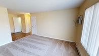 This single-floor residential space offers a straightforward and functional layout designed for comfortable everyday living. The interior is finished with warm, pale yellow walls that lend a bright and welcoming ambiance throughout. The main living area features plush wall-to-wall carpeting in a neutral beige tone, providing a soft and comfortable underfoot experience while complementing the overall color palette of the space. Clean white baseboards provide a crisp contrast against the warm walls, contributing to a tidy and well-maintained appearance. The room is equipped with standard electrical outlets and wall-mounted fixtures, reflecting practical utility throughout. A paneled white interior door leads to an adjacent area, and an open hallway connects the space to additional rooms, hinting at a multi-room layout that flows naturally from one area to the next. The ceilings are smooth and finished in white, contributing to the sense of openness within the space. The consistent use of neutral tones across walls, trim, and flooring creates a cohesive and move-in ready environment that provides a versatile backdrop for a variety of interior furnishing styles.