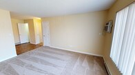 This single-floor residential space offers a straightforward and functional layout designed for comfortable everyday living. The interior is finished with warm, pale yellow walls that lend a bright and welcoming ambiance throughout. The main living area features plush wall-to-wall carpeting in a neutral beige tone, providing a soft and comfortable underfoot experience while complementing the overall color palette of the space. Clean white baseboards provide a crisp contrast against the warm walls, contributing to a tidy and well-maintained appearance. The room is equipped with standard electrical outlets and wall-mounted fixtures, reflecting practical utility throughout. A paneled white interior door leads to an adjacent area, and an open hallway connects the space to additional rooms, hinting at a multi-room layout that flows naturally from one area to the next. The ceilings are smooth and finished in white, contributing to the sense of openness within the space. The consistent use of neutral tones across walls, trim, and flooring creates a cohesive and move-in ready environment that provides a versatile backdrop for a variety of interior furnishing styles.