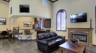 This single-story space showcases an open-concept layout defined by impressively tall ceilings that lend an expansive, airy quality to the interior. The living area is anchored by a classic wood-mantled fireplace, complemented by a dark leather sofa and rustic wood coffee table that reinforce a warm, traditional aesthetic. Stone tile flooring runs throughout, adding durability and a grounded, earthy tone to the space.

A distinctive arched window with rich dark-wood plantation shutters serves as a standout architectural feature, drawing in natural light while offering adjustable privacy. The same dark wood shutters are echoed across additional windows, ensuring a cohesive design language throughout.

Adjacent to the living area, a built-in bar or kitchenette zone features dark cabinetry, a peninsula counter with bar seating, and what appears to be a small refrigerator — ideal for entertaining. A wall-mounted television is positioned both in the upper corner of the living space and above the bar area, adding modern functionality to the otherwise traditionally styled interior. Crown molding and warm beige walls complete the inviting, cohesive atmosphere.