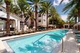 Located at 270 Courtney Lakes Cir in West Palm Beach, Florida, this two-floor property showcases a beautifully designed outdoor amenity space that evokes a true resort living experience. The architecture reflects a Mediterranean-inspired aesthetic, with crisp white stucco exteriors, terracotta tile rooflines, arched entryways, and shuttered windows that lend a timeless, elegant character throughout.

At the heart of the outdoor space is a generously sized lap pool with crystal-clear turquoise water, framed by wide tiled pool decks and lined with an array of lounge chairs — from teal to sage green — perfect for sunbathing beneath the South Florida sun. Towering date palms and lush tropical plantings create a canopy of greenery that enhances privacy and ambiance throughout the courtyard.

A covered outdoor lounge area features exposed wood-beam ceilings, decorative patterned tile accent walls, a mounted flat-screen television, and comfortable seating, serving as an ideal gathering space. A sleek water feature adds a calming focal point nearby. Private cabanas with lattice-style screens offer shaded retreats along the pool's edge.

Wide paver pathways connect the various outdoor zones, and a grand arched passageway framed by dramatic tropical foliage leads into the main building, where ceiling fans and open sightlines create an airy, welcoming entry experience.