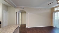 This well-appointed 1-bedroom, 1-bathroom residence is thoughtfully designed with classic architectural details that lend character and elegance throughout. The living area showcases rich, dark hardwood flooring that contrasts beautifully with the neutral, warm-toned walls, creating a welcoming and cohesive aesthetic. Crown molding trims the ceilings, adding a refined touch to the overall design. A standout architectural feature is the decorative arched doorway flanked by white column pilasters, which elegantly frames the passage between rooms and adds a sense of sophistication to the open layout. The kitchen is positioned adjacent to the living space, featuring a granite countertop that provides both functionality and style. Ceiling vents are strategically placed throughout to ensure comfortable climate control. The layout flows naturally between spaces, with the arched hallway leading toward the bedroom and bathroom. Neutral wall tones throughout create a versatile backdrop that complements a variety of interior styles. The combination of classic detailing, quality finishes, and an open, connected floor plan makes this a comfortable and stylish single-floor residence.