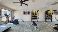This fitness center is a comprehensively outfitted single-floor exercise facility designed to support a wide range of workout routines. The space is finished with a distinctive modular carpet in a grey and green geometric pattern, lending the room a modern, energetic aesthetic that complements its function. Recessed lighting and a ceiling fan ensure the space remains well-lit and comfortable during use.

The cardio zone is positioned along the windowed wall, where oversized square windows frame direct views of an outdoor pool deck complete with lounge seating and vibrant red umbrellas. This area houses treadmills, a stationary bike, and an elliptical trainer, allowing users to exercise while enjoying natural light and outdoor scenery.

On the opposite side of the room, a dedicated strength and functional training area features a full dumbbell rack with a wide range of weights, an adjustable workout bench, and a multi-purpose training rig equipped with TRX suspension straps, medicine balls, and resistance accessories. A large floor-to-ceiling mirror spans one wall, enhancing the sense of space and aiding in form correction during exercise.

A wall-mounted flat-screen TV adds an entertainment element, while posted fitness center guidelines ensure a structured and safe environment. The facility offers direct access to the outdoor pool area, seamlessly connecting indoor exercise with outdoor amenities.