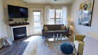 The Clark's 1-bedroom, 1-bathroom floor plan offers a thoughtfully designed single-level layout finished with dark hardwood-style flooring that runs consistently throughout the home. The living room serves as the central gathering space, anchored by a corner fireplace with a wood mantel and complemented by a wall-mounted television. A glass-top coffee table and a direct-access door lead to the private balcony, while large windows dressed in sheer white curtains invite abundant natural light and views of the surrounding neighborhood.

The kitchen is well-equipped with crisp white cabinetry extending to the upper walls, providing generous storage. A mosaic tile backsplash adds texture, while light-toned countertops and stainless steel appliances — including a range, over-the-range microwave, and dishwasher — complete the functional workspace. Under-cabinet lighting enhances visibility and ambiance.

The private balcony extends the living space outdoors, offering open views of the residential community and enough room for seating. The overall aesthetic throughout the residence is modern and cohesive, combining neutral tones with contemporary fixtures for a clean, inviting atmosphere.
