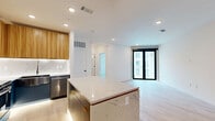 Located at 1923 Broadway Blvd in Kansas City, MO, this thoughtfully designed one-bedroom, one-bathroom residence offers a well-appointed layout across a single floor. The kitchen is an immediate standout, featuring bold teal appliances — including a refrigerator and range — paired with warm wood cabinetry, under-cabinet lighting, and a striking center island with a marble-look quartz countertop and teal base paneling. Light hardwood flooring ties the kitchen and living areas together, creating a bright, cohesive feel. The living area offers a generous open canvas with light walls and ample space for flexible furniture arrangements. The bedroom is a comfortable retreat, finished with plush carpeting, a ceiling fan, and a large window with black-framed trim that allows natural light while providing views of the surrounding urban streetscape. The bathroom is notably elegant, showcasing bold navy vertical tile in the glass-enclosed shower, marble-patterned tile flooring, a sleek floating vanity with gold hardware, and matte black fixtures — a refined contrast of modern and luxurious elements. A dedicated closet and hallway round out the functional layout of this well-designed urban residence.