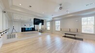 Located at 1810 Arden Creek Pl in Charlottesville, VA, this purpose-built fitness and dance studio offers a well-appointed, single-room space designed for movement, exercise, and instruction. The studio features light-toned hardwood flooring throughout, providing a clean, durable surface ideal for a variety of fitness activities. A full-length mirrored wall spans one side of the room, enhancing the sense of openness while offering essential visual feedback for dancers and exercisers alike. Wall-mounted ballet barres extend along multiple walls, supporting barre workouts, stretching, and dance training. A wall-mounted flat-screen television serves as an instructional display, while recessed lighting and an overhead ceiling fan ensure a comfortable and well-lit environment. Natural light filters in through multiple windows fitted with plantation shutters, adding brightness and a connection to the outdoors. A flat workout bench paired with an exercise mat provides space for strength and conditioning exercises. Built-in dark cubby shelving units offer organized storage for yoga mats, foam rollers, resistance bands, and dumbbells, keeping equipment neatly accessible. Crown molding and a tray ceiling add a refined architectural touch, elevating the overall aesthetic of this thoughtfully designed studio space.
