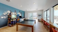This single-floor game room in Aurora, Colorado, is a thoughtfully designed recreational space that balances energetic entertainment with comfortable gathering areas. The room centers around a full-size Brunswick ping-pong table positioned on light wood-toned flooring, providing ample space for play and movement throughout.

One of the most striking design elements is a bold, teal-toned nature mural spanning an entire accent wall, depicting silhouetted pine trees and mountain scenery. This artistic feature anchors the entertainment corner of the room, where a collection of classic stand-up arcade machines adds a retro gaming experience. Adjacent to the arcade units, a built-in wet bar with dark countertops, under-counter refrigeration, and a sink offers convenient beverage service.

On the opposite side, a full brick accent wall provides textural contrast and hosts an oversized wall-mounted Scrabble board, adding a social board game option to the space. A window-lined wall with a built-in bar-height counter and red bar stools offers additional seating with panoramic outdoor views. Large sliding glass doors lead directly to an outdoor terrace with seating, extending the usable social space and flooding the interior with natural light.