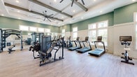Located at 3730 Dupont Avenue in Jacksonville, FL, this fitness center offers a generously sized, single-floor exercise space designed with both functionality and aesthetic appeal in mind. The main workout area is defined by soaring ceilings adorned with rustic exposed wood beams and coffered detailing, creating an open, airy atmosphere that enhances the sense of space throughout.

Large, multi-pane windows line the walls, flooding the interior with abundant natural light and providing views of the surrounding exterior. The light wood-tone flooring complements the soft sage green and crisp white wall paneling, giving the space a clean, welcoming ambiance.

Oversized ceiling fans are strategically positioned to ensure comfortable airflow across the entire floor. The main exercise area is well-appointed with a comprehensive range of equipment, including multiple treadmills, elliptical machines, and multi-station strength training systems. A dedicated free weights section features kettlebell racks, adjustable benches, and a cable machine, while a separate area is outfitted with medicine balls, stability equipment, and floor space for functional training. Full-length mirrors throughout amplify the brightness and allow users to monitor their form. A compact utility station near the entrance adds convenience to the overall layout.
