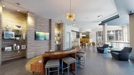 Located at 616 Oberlin Dr in Raleigh, NC, this single-floor communal amenity space showcases a sophisticated, contemporary design that blends style and functionality. The entry area features large-format tile flooring, textured stacked-stone accent walls, and floor-to-ceiling glass partitions that create a bright, open atmosphere while defining distinct zones within the space.

At the heart of the lobby is a curved wooden reception bar with upholstered stools, anchored beneath a striking gold sputnik chandelier—a standout design element that sets a modern, upscale tone. Teal-painted ceiling accents contrast elegantly with crisp white walls throughout, adding depth and visual interest.

A glass-enclosed lounge area offers an intimate seating space with a bold blue velvet sofa, while the open main lounge features a curved dark sectional, contemporary armchairs, and coordinating side tables. A café-style nook with a yellow tufted booth and natural wood cabinetry provides a casual gathering spot, complemented by additional seating clusters arranged throughout.

Floor-to-ceiling windows along one side of the space flood the interior with natural light and offer views of the surrounding courtyard and residential buildings. The overall ambiance is polished and welcoming, combining mid-century modern influences with current design sensibilities to create an inviting, multi-functional community space.