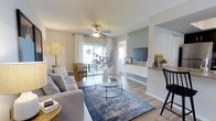 This single-floor, 2-bedroom, 2-bathroom residence offers a thoughtfully designed layout that balances comfort and functionality. The living area serves as the heart of the home, featuring light-toned luxury vinyl plank flooring that runs throughout, creating a cohesive and clean aesthetic. A ceiling fan with integrated lighting centers the room, while a large sliding glass door floods the space with natural light and provides access to an outdoor area, offering pleasant views and an airy atmosphere. Neutral wall tones are complemented by a soft sage accent wall that frames a built-in media niche, adding a subtle architectural detail to the living space. An open-concept design connects the living area to a dedicated workspace or dining bar, complete with a countertop ledge and bar seating — ideal for everyday convenience. White sheer curtains frame the windows, softening the natural light and enhancing the bright, welcoming ambiance. The overall design palette emphasizes calm, neutral tones with modern furnishing potential, making the space feel both versatile and inviting. With two full bathrooms and two bedrooms, this residence provides practical accommodations suited for a range of lifestyles.