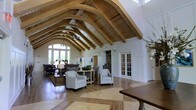 This remarkable single-floor space makes an immediate impression with its dramatic vaulted ceiling, defined by a series of arched wooden beams that span the length of the room in a graceful, cathedral-like fashion. The natural wood tones of the ceiling structure contrast beautifully against crisp white walls, creating a warm yet refined aesthetic. The flooring features an elegant inlaid pattern where rich hardwood meets lighter stone-toned tiles, adding visual interest and grounding the space with a sense of understated luxury. Large windows at the far end of the main gathering area flood the interior with natural light, framing views of the surrounding greenery and enhancing the open, airy feel of the room. The main seating area is furnished with classic upholstered armchairs and sofas arranged in a welcoming layout, ideal for conversation and relaxation. French doors with white millwork paneling lead to adjacent rooms, suggesting a thoughtful, well-connected floor plan. A chandelier adds a subtle decorative touch beneath the soaring ceiling. The overall ambiance is one of elegant comfort — sophisticated in its architectural detail, yet approachable and inviting in its design and furnishings.