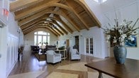 This remarkable single-floor space makes an immediate impression with its dramatic vaulted ceiling, defined by a series of arched wooden beams that span the length of the room in a graceful, cathedral-like fashion. The natural wood tones of the ceiling structure contrast beautifully against crisp white walls, creating a warm yet refined aesthetic. The flooring features an elegant inlaid pattern where rich hardwood meets lighter stone-toned tiles, adding visual interest and grounding the space with a sense of understated luxury. Large windows at the far end of the main gathering area flood the interior with natural light, framing views of the surrounding greenery and enhancing the open, airy feel of the room. The main seating area is furnished with classic upholstered armchairs and sofas arranged in a welcoming layout, ideal for conversation and relaxation. French doors with white millwork paneling lead to adjacent rooms, suggesting a thoughtful, well-connected floor plan. A chandelier adds a subtle decorative touch beneath the soaring ceiling. The overall ambiance is one of elegant comfort — sophisticated in its architectural detail, yet approachable and inviting in its design and furnishings.
