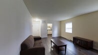 This single-floor space offers a practical and well-organized layout designed for everyday living. The living area serves as the central hub of the unit, featuring rich dark hardwood flooring that contrasts cleanly against the neutral, light-toned walls and bright white trim. A window along one wall allows natural light to filter into the room, contributing to an open and airy atmosphere. The living space provides ample room for standard furnishings, as evidenced by the comfortable seating and coffee table arrangement visible within. A clearly defined entryway with a white front door provides a clean point of entry, complete with overhead ventilation and a ceiling-mounted light fixture. Just off the main living area, an open doorway leads directly into the kitchen, which features light wood cabinetry, a countertop workspace, and essential appliances. The seamless flow between the living and kitchen areas enhances the overall sense of connectivity within the unit. Baseboard heating is installed along the perimeter walls, ensuring comfort throughout the cooler months. The overall ambiance is clean, neutral, and functional, making the space well-suited for a variety of uses and easily adaptable to different furnishing styles.