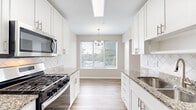 This well-appointed single-floor residence in Rockville, MD offers four bedrooms and four full bathrooms across a generously sized floor plan. The bright living room features light hardwood-style flooring, a vaulted ceiling, and a bank of large windows that flood the space with natural light and offer views of the surrounding greenery. The kitchen is thoughtfully designed with white shaker cabinetry, granite countertops, a diamond-pattern tile backsplash, stainless steel appliances including a gas range, and a chandelier above a dining nook framed by an outdoor-facing window. The spacious primary bedroom is finished with plush carpeting and crown molding, and connects to a luxurious en-suite bathroom featuring a soaking tub with gold fixtures and marble-look tile flooring. The primary suite also includes a large walk-in closet outfitted with built-in shelving, hanging rods, and drawer storage. Additional bedrooms offer carpeted or hardwood-style flooring, crown molding, and ample closet space with wire shelving. A dedicated in-unit laundry area houses a full-size washer and dryer. Neutral tones and clean finishes throughout create a bright, cohesive ambiance.