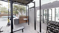 This outdoor amenity space offers a well-rounded recreational experience set within a contemporary building courtyard. The deck is finished with a smooth grey surface that defines distinct activity zones, providing ample room for movement and leisure. A vibrant blue STIGA ping-pong table serves as a central focal point for casual recreation, positioned in the open courtyard area alongside raised wooden planter boxes that introduce natural greenery, including a mature olive tree that adds character to the setting. Adjacent to the open play area, a dedicated outdoor fitness zone is sheltered beneath a sturdy black metal canopy structure. This covered workout area is equipped with a multi-functional power rack, pull-up bars, adjustable bench, and additional strength training equipment, with large wall-mounted mirrors enhancing the sense of space. The grey tile flooring in the fitness zone provides a durable, weather-resistant surface suited for exercise. The surrounding building façade features large windows and private balconies on upper floors, creating an enclosed yet open-air atmosphere. A glimpse of a pool area is also visible in the background, suggesting this outdoor deck is part of a broader amenity offering within the property.