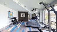 This fitness center offers a thoughtfully designed workout space that combines functionality with contemporary style. The room features a striking striped floor in varying shades of blue, gray, and white, lending the space a dynamic, energetic atmosphere. The walls are finished in crisp white, with one accent wall in deep charcoal gray serving as a bold focal point, adorned with a framed geometric artwork that adds an artistic touch to the environment.

The cardio zone is well-equipped with multiple treadmills arranged in a row, complemented by additional cardiovascular machines toward the rear of the space. A dedicated free weights area lines one wall, featuring a fully stocked dumbbell rack with a flat bench for strength training exercises. Overhead, ceiling-mounted fans promote air circulation throughout the facility.

Storage cubbies are built into the back wall, providing convenient organization for personal belongings. Natural light filters in through windows along one side, brightening the space and creating an inviting atmosphere. The ceiling incorporates recessed lighting alongside track lighting, ensuring the room remains well-lit at all hours. The overall layout promotes efficient use of space while maintaining an open, comfortable environment for exercising.