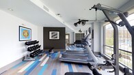 This fitness center offers a thoughtfully designed workout space that combines functionality with contemporary style. The room features a striking striped floor in varying shades of blue, gray, and white, lending the space a dynamic, energetic atmosphere. The walls are finished in crisp white, with one accent wall in deep charcoal gray serving as a bold focal point, adorned with a framed geometric artwork that adds an artistic touch to the environment.

The cardio zone is well-equipped with multiple treadmills arranged in a row, complemented by additional cardiovascular machines toward the rear of the space. A dedicated free weights area lines one wall, featuring a fully stocked dumbbell rack with a flat bench for strength training exercises. Overhead, ceiling-mounted fans promote air circulation throughout the facility.

Storage cubbies are built into the back wall, providing convenient organization for personal belongings. Natural light filters in through windows along one side, brightening the space and creating an inviting atmosphere. The ceiling incorporates recessed lighting alongside track lighting, ensuring the room remains well-lit at all hours. The overall layout promotes efficient use of space while maintaining an open, comfortable environment for exercising.