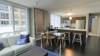 This modern single-floor residence offers a thoughtfully designed open-concept layout that seamlessly integrates the living, dining, and kitchen areas into one cohesive and functional space. Dark hardwood flooring runs throughout, providing a rich, warm contrast to the neutral grey walls and crisp white accents. The living area is anchored by a comfortable sofa adorned with deep blue accent pillows and a vibrant area rug, creating an inviting atmosphere. Large windows dressed with vertical blinds allow abundant natural light to fill the space while offering views of the surrounding exterior. A striking large-scale framed artwork adds a sophisticated focal point to the living zone. The dining area features a sleek glass-top table with modern wooden chairs, positioned to take full advantage of the open flow. The kitchen is well-appointed with dark cabinetry, stainless steel appliances including a built-in microwave and range, and a spacious island with bar-height seating — ideal for casual dining or entertaining. Pendant lighting above the island adds a warm, stylish touch. The overall ambiance is contemporary and polished, balancing clean architectural lines with carefully chosen furnishings for a refined yet comfortable living experience.