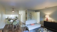 This single-floor residence offers a thoughtfully designed open-concept layout that integrates the living, dining, and kitchen areas into one cohesive and functional space. The living area serves as the heart of the home, featuring rich hardwood flooring, neutral-toned walls accented by a bold gray feature wall, and built-in shelving that adds both character and practicality. A ceiling fan overhead enhances comfort and airflow throughout the space. The kitchen, visible from the living and dining areas, is equipped with modern appliances including a full-size refrigerator, ample cabinetry in a clean white finish, and a countertop workspace ideal for meal preparation. A casual dining area with a round glass table and contemporary seating sits at the intersection of the kitchen and living room, making it perfect for everyday meals or entertaining. Natural light filters in from nearby windows, brightening the entire space. The open doorway visible from the main living area suggests additional private rooms beyond, contributing to an overall sense of spaciousness and flow. The combination of modern décor, warm flooring, and a neutral color palette creates an inviting and livable ambiance throughout.